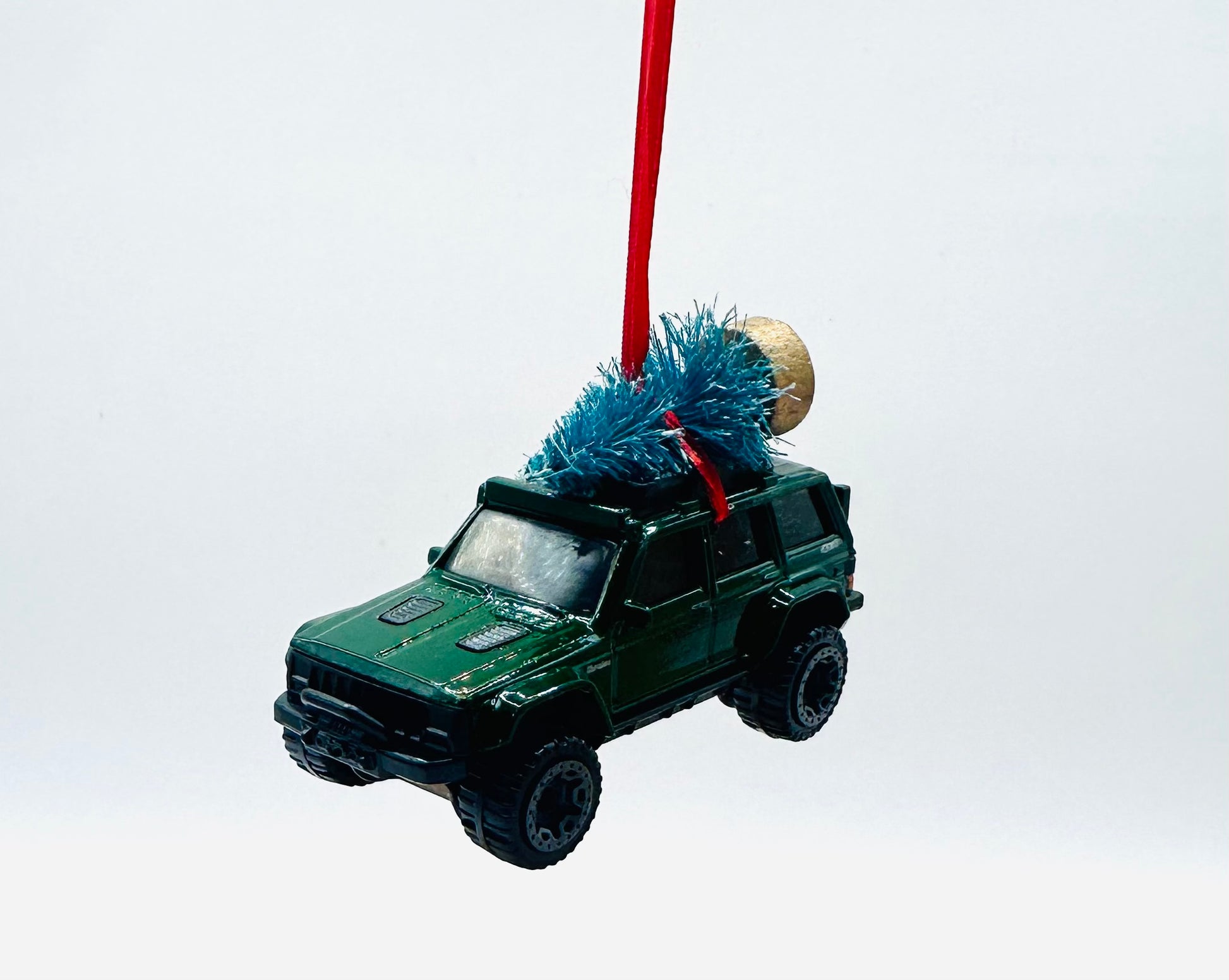 Green toy Jeep Cherokee with a Christmas tree on top, hanging from a red string against a white background.
